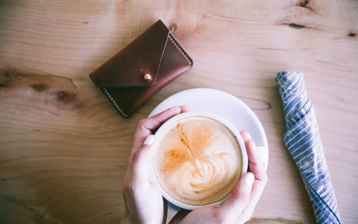 Cliente tenant un cappuccino sur une table en bois clair, à côté d’un portefeuille.